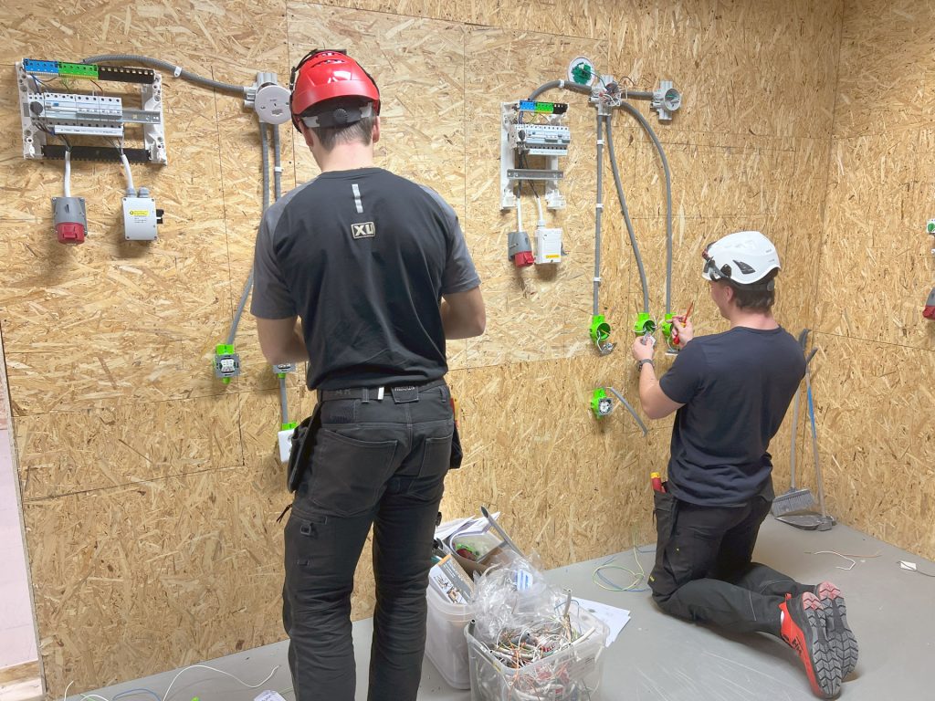 Två personer tränar praktiskt på elinstallation i en utbildningsmiljö med träväggar, skyddshjälmar och arbetskläder. De monterar elcentraler, kopplingsdosor och eluttag med noggrannhet. Här får deltagarna den yrkeskunskap som krävs – för riktiga jobb kräver riktig kompetens.