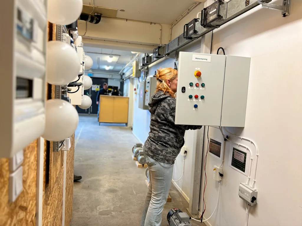 En elev arbetar med en stor elstyrningspanel som är monterad på väggen. Panelen har flera knappar och vred, och det verkar vara ett moment inom elteknik eller automation. Bilden är tagen i en utbildningsmiljö för Nationell yrkesutbildning i Göteborg, där elever får träna praktiskt på tekniska installationer. I bakgrunden syns en lång korridor med fler installationer längs väggarna.