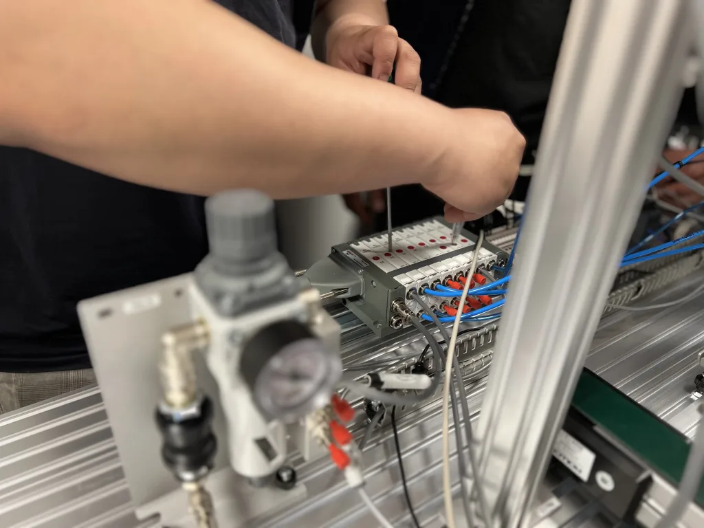 Närbild på en person som arbetar med en pneumatisk styrmodul. Personen använder en skruvmejsel för att montera eller justera komponenter. Modulen har flera blå slangar anslutna och är monterad på en aluminiumram. I förgrunden syns en tryckregulator och en tryckmätare i metall, och runt om finns en ren och tekniskt organiserad arbetsmiljö.