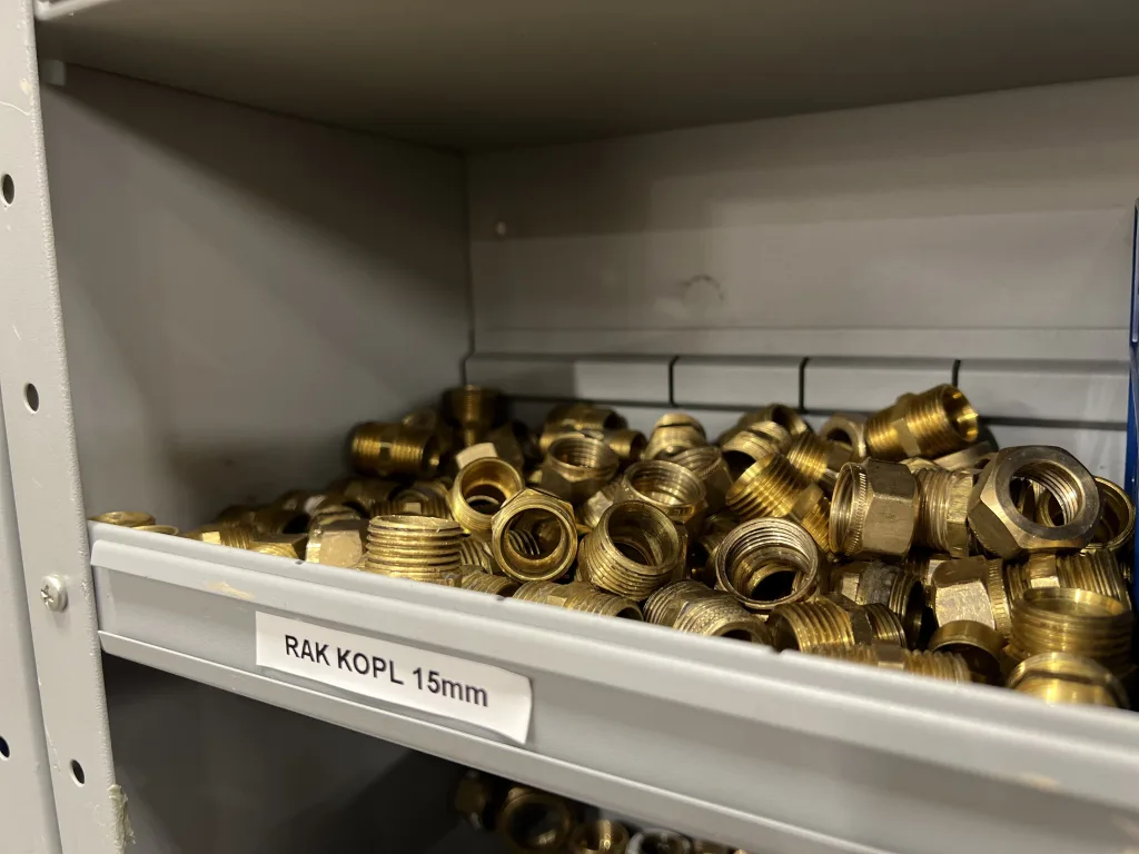 Hyllplan i metall med märkning "RAK KOPL 15mm", fylld med mässingskopplingar i olika storlekar och former. De gängade kopplingarna är välorganiserade i hyllor för enkel åtkomst och installation.