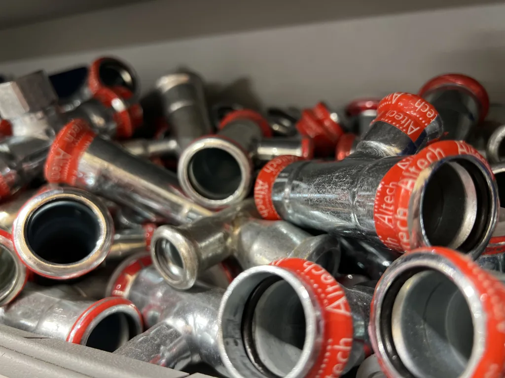 Närbilder på ett lager med rörkopplingar i metall från varumärket Altech. Kopplingarna är försedda med röda ringar som visar Altech-logotypen. De är prydligt organiserade i en låda och redo att användas för installation i olika rörsystem.