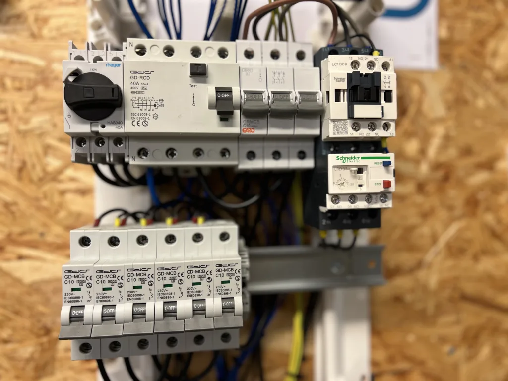 Närbilder på en elcentral monterad på en träpanelvägg. Den innehåller flera automatsäkringar (GD-MCB CE C10), en jordfelsbrytare (GD-RCD 40A) och kontaktorer från märkena Schneider Electric och Hager. Flera blå och gula kablar är inkopplade i modulerna. Bilderna visar både helhetsvy och närbilder på olika komponenter i elcentralen, som används för att säkra och styra elektriska system.