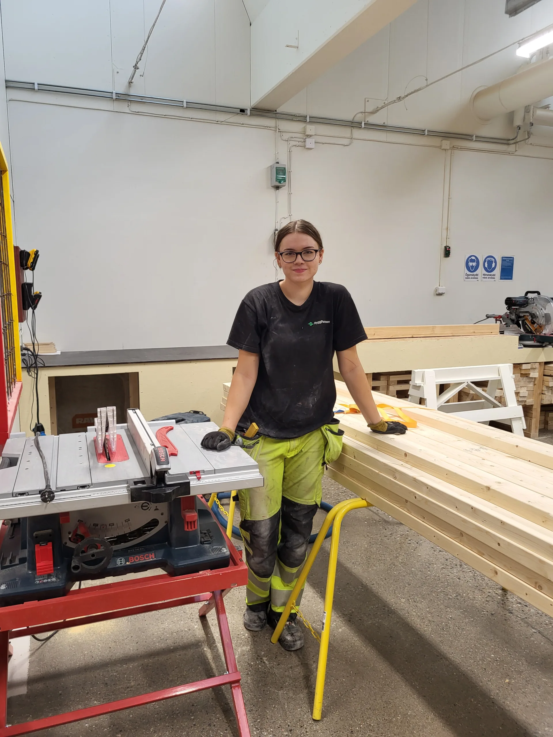En ung kvinna i arbetskläder står vid en bordssåg i en träverkstad, omgiven av plankor och verktyg. Hon bär glasögon och handskar, vilket visar fokus på säkerhet och yrkesskicklighet. Eskilstuna byggnadsträarbetare