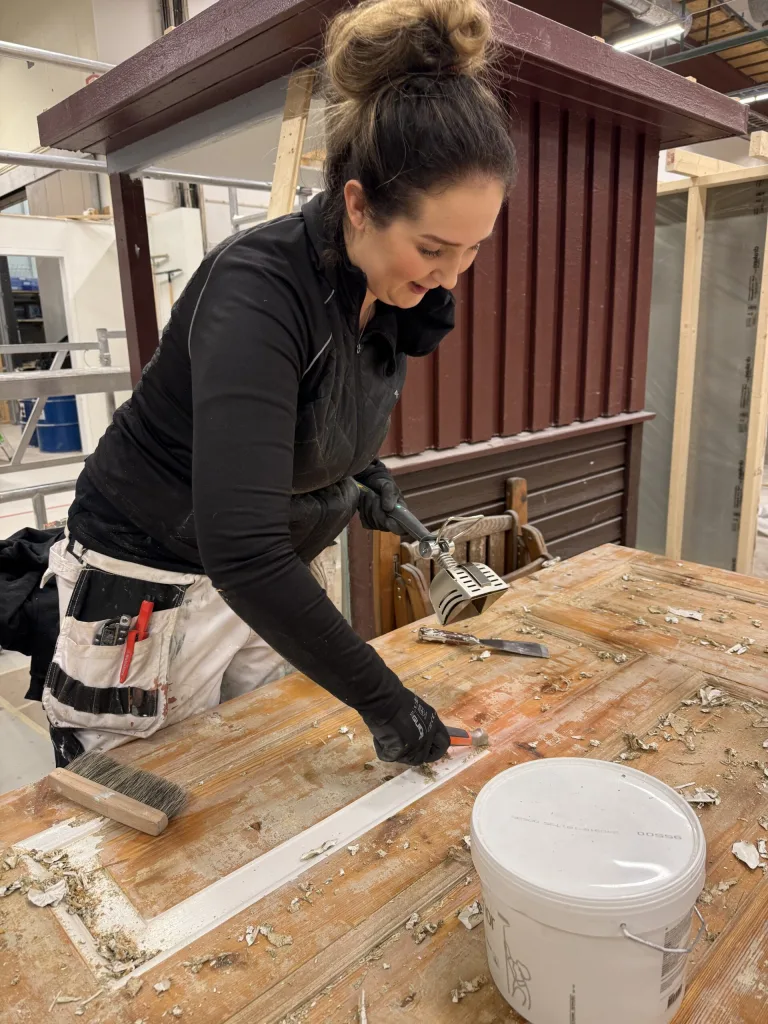 En kvinna arbetar med att renovera en träyta. Hon använder olika handverktyg, inklusive en skrapa, för att ta bort gammal färg från ett träbord. Hon bär arbetskläder med fickor och penslar, och ser koncentrerad och engagerad ut. I bakgrunden syns en byggarbetsplats med träkonstruktioner och en röd fasad. En vit plasthink står på bordet som används för att samla bortskrapat material.