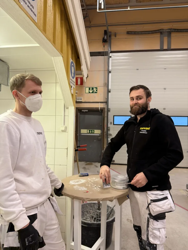 Två män arbetar tillsammans vid ett runt bord täckt med brunt papp i en verkstadsmiljö under en utbildning i Halmstad. En av dem bär vitt målarställ och munskydd, medan den andra har en svart tröja med Movant-logotyp och vitt arbetsställ. De verkar arbeta med spackel eller slipning, och bordet är täckt av damm och verktyg. I bakgrunden syns en verkstadsdörr och skyltar med säkerhetsanvisningar. Båda är fokuserade och samarbetar aktivt i det praktiska momentet.
