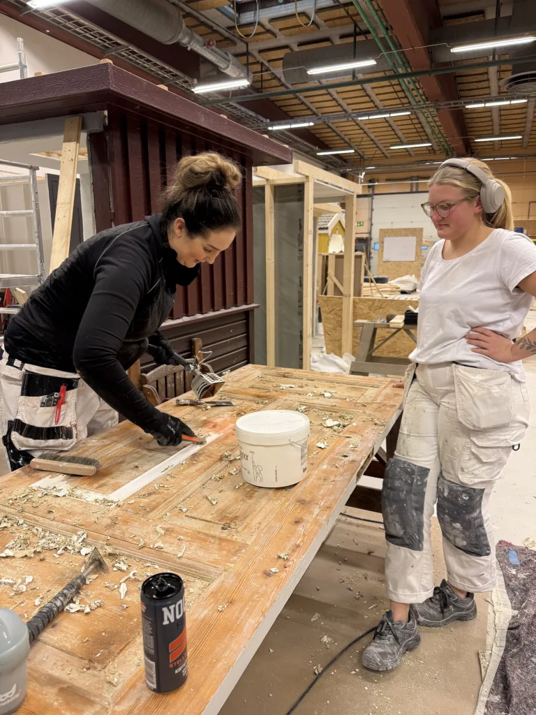 Två kvinnor arbetar tillsammans i en verkstadsmiljö hos Movant i Halmstad. En av dem, med mörkt hår i knut och svart tröja, skrapar bort gammal färg från en träyta på ett stort bord. Den andra kvinnan, med ljust hår, hörlurar och glasögon, står bredvid och tittar på. Båda bär arbetskläder, och bordet är täckt med färgflagor och verktyg. I bakgrunden syns en röd fasad, ett arbetsområde och byggmaterial.