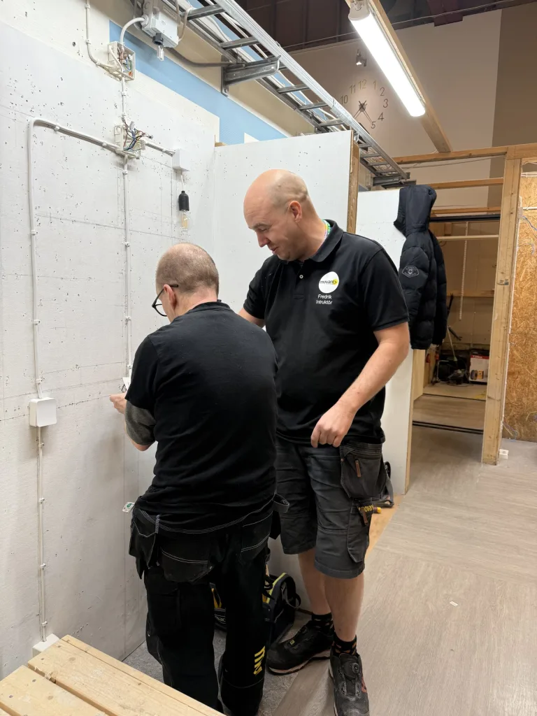 Två män i svarta arbetskläder arbetar med en elinstallation i en lärandemiljö. Den ena mannen bär en pikétröja med texten "Movant Fredrik Instruktör" och instruerar den andra mannen som monterar elkablar på en vit vägg. På väggen syns flera kablar, eluttag och kopplingsdosor. Miljön är en verkstad med träväggar och takbelysning.