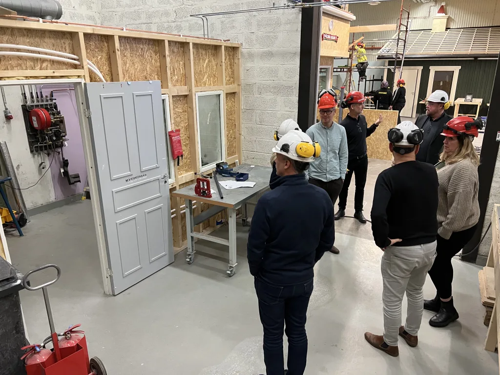 En grupp människor i arbetskläder och skyddshjälmar samlas i en verkstadsmiljö. De verkar ha en genomgång eller diskussion om projektet. I bakgrunden syns en byggnadskonstruktion med dörr och fönster installerade, och en person som arbetar på en byggställning. Golvet är rent och betonggrått och verkstaden är full av byggmaterial och verktyg. På en vägg hänger ett brandlarm och en brandslang.