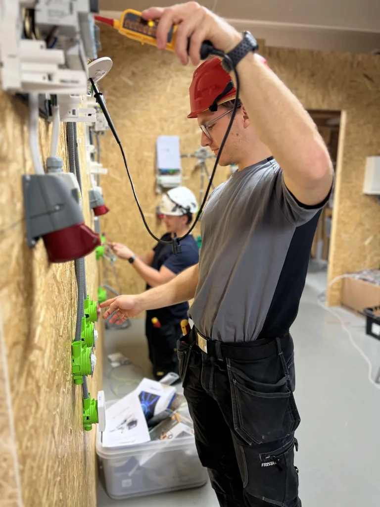Två personer arbetar med elinstallationer på en vägg täckt med OSB-skivor. Den främre personen, med röd skyddshjälm och grå-svart t-shirt, använder en spänningsprovare för att mäta eller kontrollera elanslutningar. Han har ett verktygsbälte med olika verktyg. Den andra personen, längre bak, sitter på knä och arbetar också med elinstallationer. På golvet står en låda med diverse kablar och installationsmaterial.