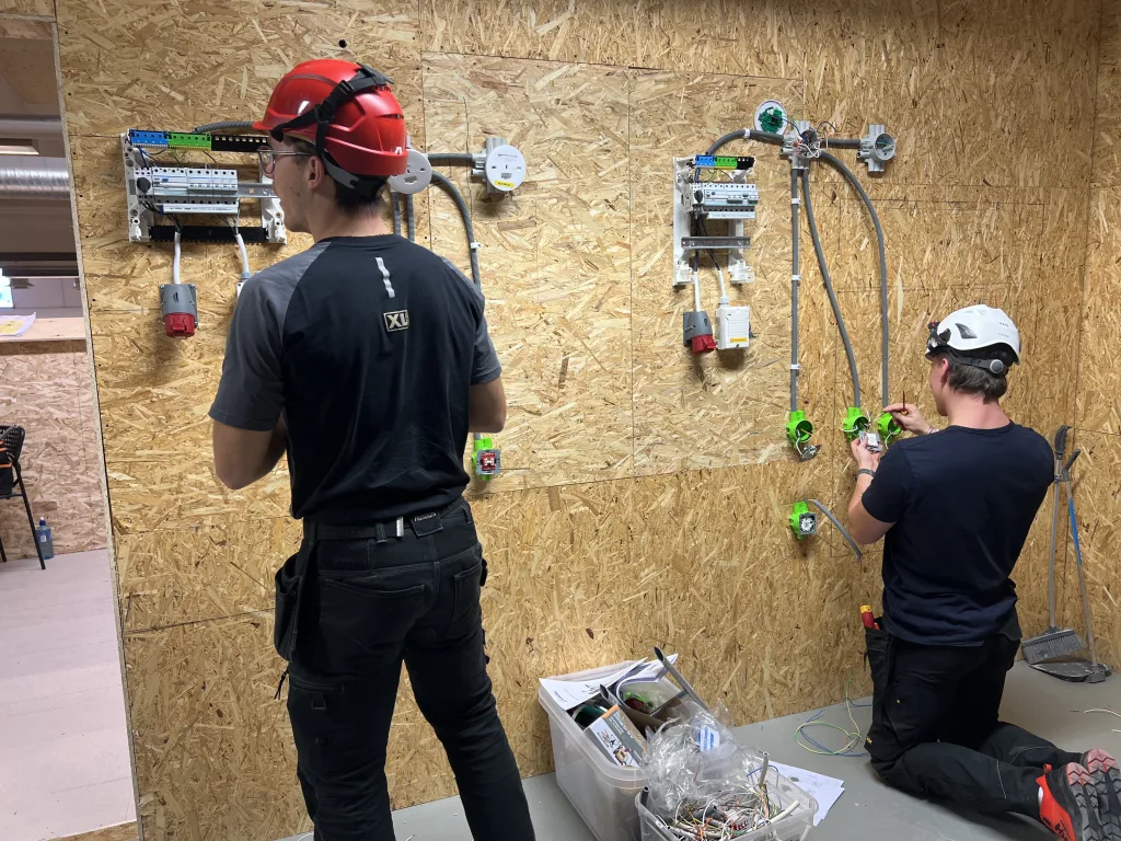 Två personer arbetar med elinstallationer på en trävägg täckt med OSB-skivor i en verkstadsmiljö. Den ena personen står med röd skyddshjälm och arbetar med en elcentral monterad på väggen. Den andra personen, med vit hjälm och hörselskydd, sitter på knä och installerar gröna väggdosor. Golvet är belamrat med dokument, kablar och installationsmaterial.