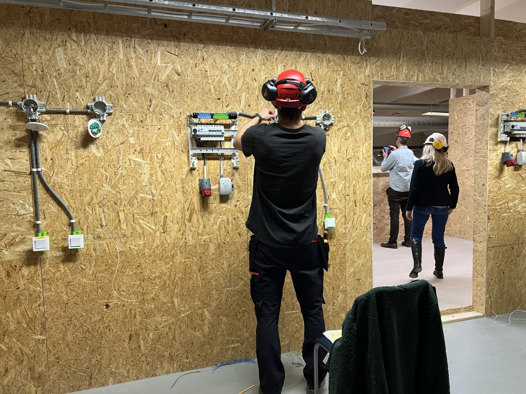 I en verkstad med väggar av OSB-skivor arbetar en person i röd hjälm och hörselskydd med elinstallationer. På väggen sitter ett elskåp och flera rördragningar för kablar. I bakgrunden syns två personer i hjälm och arbetskläder som rör sig i lokalen, vilket ger en livlig och arbetsam atmosfär. På golvet ligger en grön jacka över en stol och några verktyg syns i närheten.