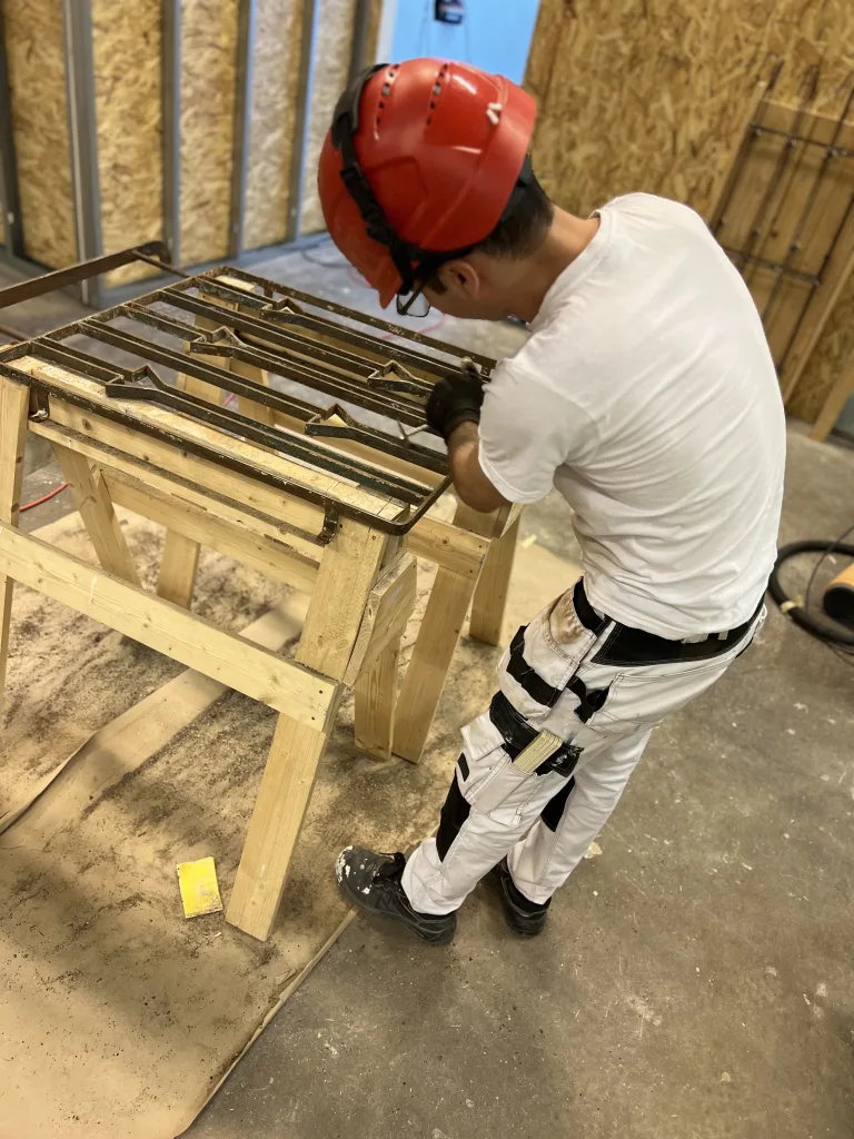 En person med röd skyddshjälm och vit t-shirt arbetar med ett metallgaller som ligger på en bock i en verkstadsmiljö. Han verkar slipa eller förbereda metallen för fortsatt arbete. Golvet är täckt med papp för att skydda det, och personen bär arbetsbyxor med verktygsfickor och arbetsskor. Bakom honom syns en vägg av OSB-skivor och några metallstänger står lutade mot väggen.