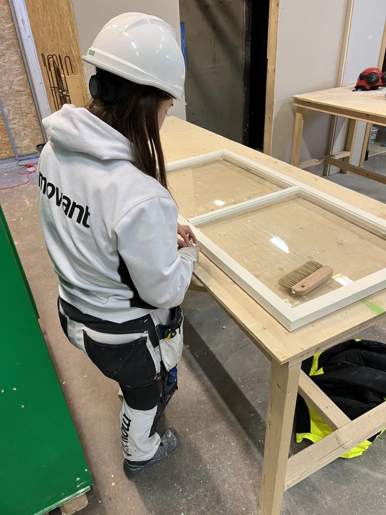 En kvinna med långt hår, vit hjälm och vit arbetsjacka med texten "movant" arbetar med ett fönsterkarm i en verkstad. Hon står vid ett arbetsbord och verkar noggrant justera eller slipa på fönsterkarmen. På bordet ligger en borste och bakom henne syns verktyg och en jacka på golvet. Verkstadsmiljön är uppbyggd i trä och OSB-skivor.