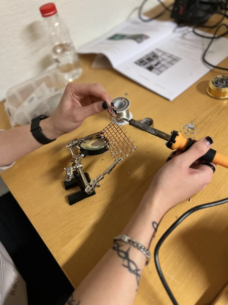 En person sitter vid ett träbord och arbetar med lödning. Personen håller i en lödkolv och ett litet rutnät i metall, som fixeras i ett litet stativ. I bakgrunden syns en vattenflaska och en öppen instruktionsbok med bilder och text. På bordet finns också en liten plastlåda med komponenter och en rulle lödtenn. Personen bär ett armband och har tatueringar på armen, vilket ger en personlig prägel i det tekniska arbetet.