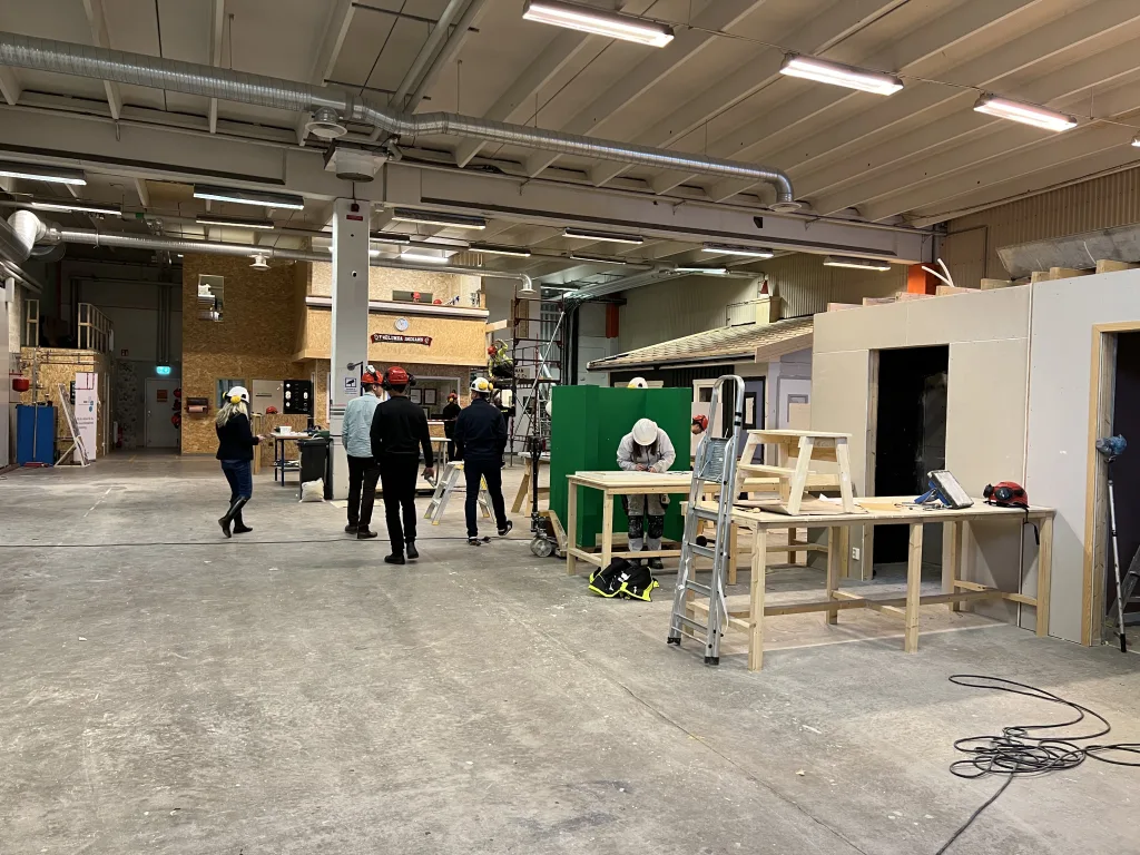 En stor och öppen verkstadsmiljö med högt i tak och flera personer som arbetar eller diskuterar. På golvet ligger arbetsmaterial och en stege står lutad mot ett arbetsbord. I förgrunden arbetar en person i vit hjälm och arbetskläder med en träkonstruktion. I bakgrunden syns OSB-beklädda rum, byggmodeller och byggställningar. Verkstaden har en blandning av arbetsbänkar, färdiga moduler och maskiner. Ljuset kommer från lysrör i taket.