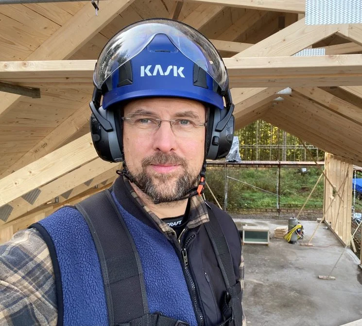 En träarbetare med skyddshjälm och hörselskydd poserar framför ett träbjälklag under uppförande. Han bär skyddsväst och flanellskjorta, med en bakgrund som visar en skog och byggställningar.