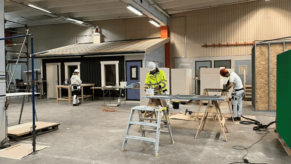 I Movants lokaler i Eskilstuna arbetar flera personer med praktiska moment. En person i varselkläder målar en planka, en annan sågar en träskiva och en tredje står vid en arbetsbänk. Lokalen är rymlig och ljus.
