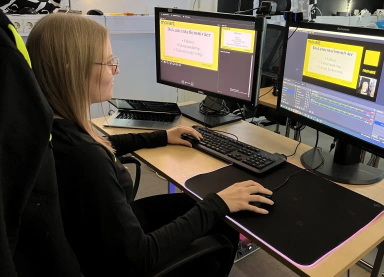 En lärare i Lund för nätverkstekniker arbetar fokuserat vid dubbla bildskärmar i ett modernt IT-klassrum. På skärmarna visas en digital presentation med teknisk dokumentation. Miljön speglar en professionell undervisningssituation inom nätverksteknik på Movant.