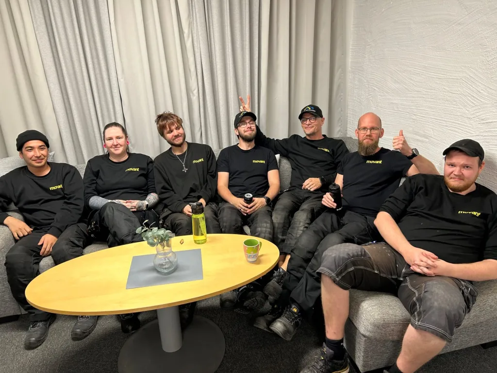 En grupp glada elever från Movant i **Karlstad** sitter tillsammans i en soffa, klädda i svarta arbetskläder med skolans logotyp. De ser ut att trivas, med leenden och avslappnad stämning. Bakgrunden består av ljusa gardiner och en vit vägg.
