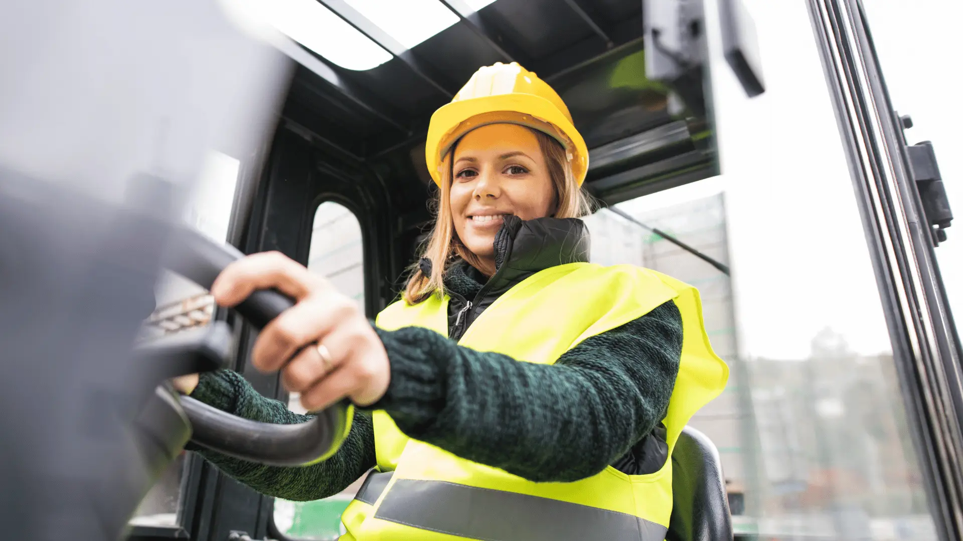 En kvinna iförd reflexväst och skyddshjälm kör en truck i en lager- eller terminalmiljö. Hon ser fokuserad och positiv ut, vilket speglar en professionell inställning till sitt arbete. Bilden representerar yrkesrollen lager-och-terminalarbetare komux