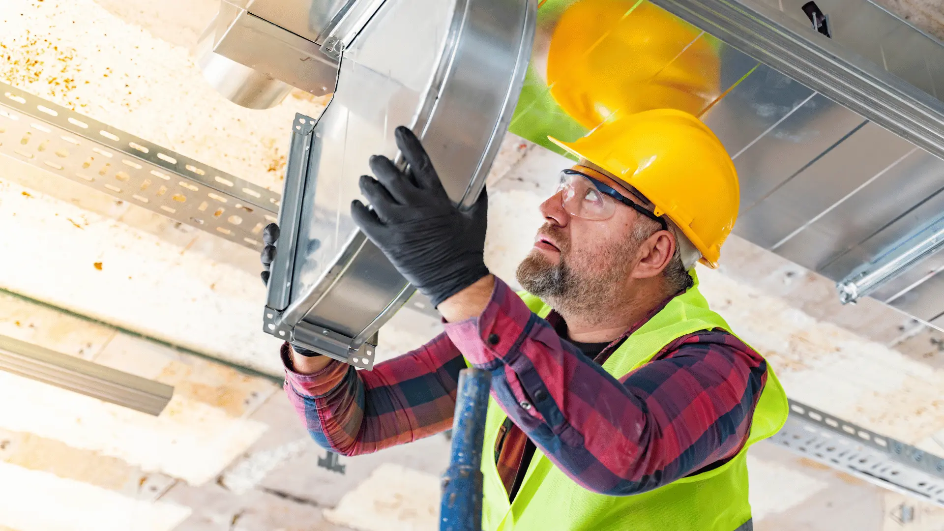 Ventilationsmontör på stege monterar ett runt ventilationsdon i taket. Han bär skyddsglasögon, hjälm och reflexväst och arbetar med precision i ett undertakssystem.