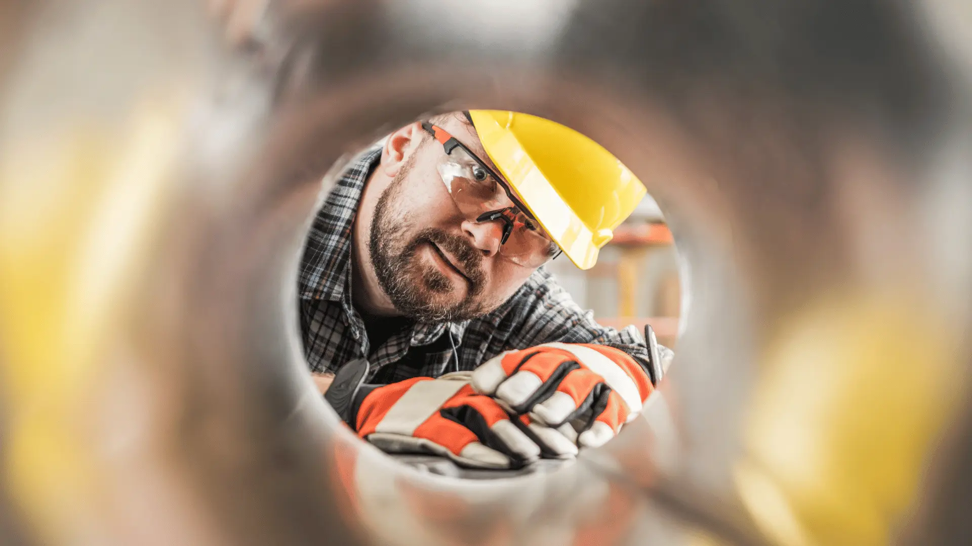 Ventilationsmontör tittar koncentrerat in i ett metallrör, sett ur ett kreativt perspektiv inifrån röret. Skyddshjälm och handskar visar på säkerhetsmedvetet arbete i verkstad eller på bygge.
