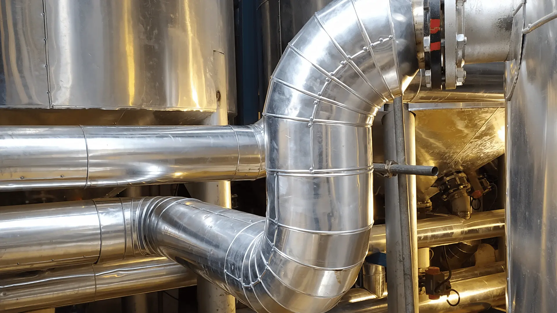 Blankpolerade ventilationsrör och kanaler i metall löper genom en industriell anläggning. Installationen visar noggrant utförda böjar och skarvar, typiskt arbete för en ventilationstekniker som installerar eller underhåller ventilationssystem.
