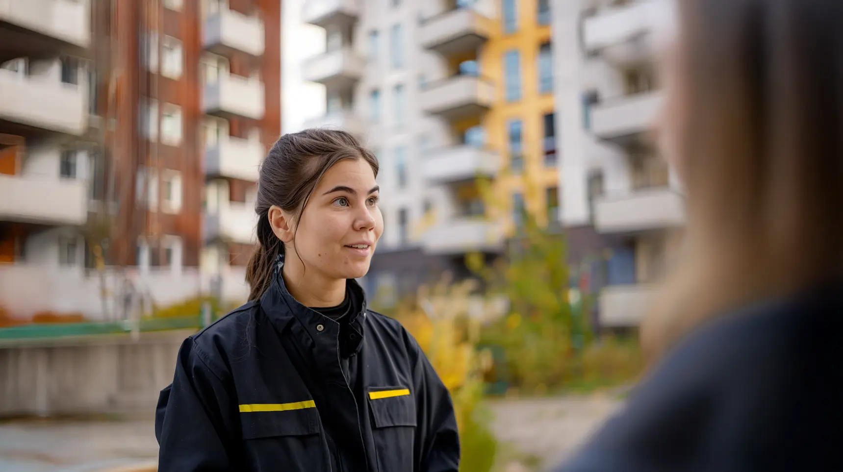 En kvinnlig fastighetsvärd i mörk arbetsjacka samtalar utomhus, med moderna flerbostadshus i bakgrunden. Hon ser engagerad ut och bidrar till en trygg och välskött boendemiljö.