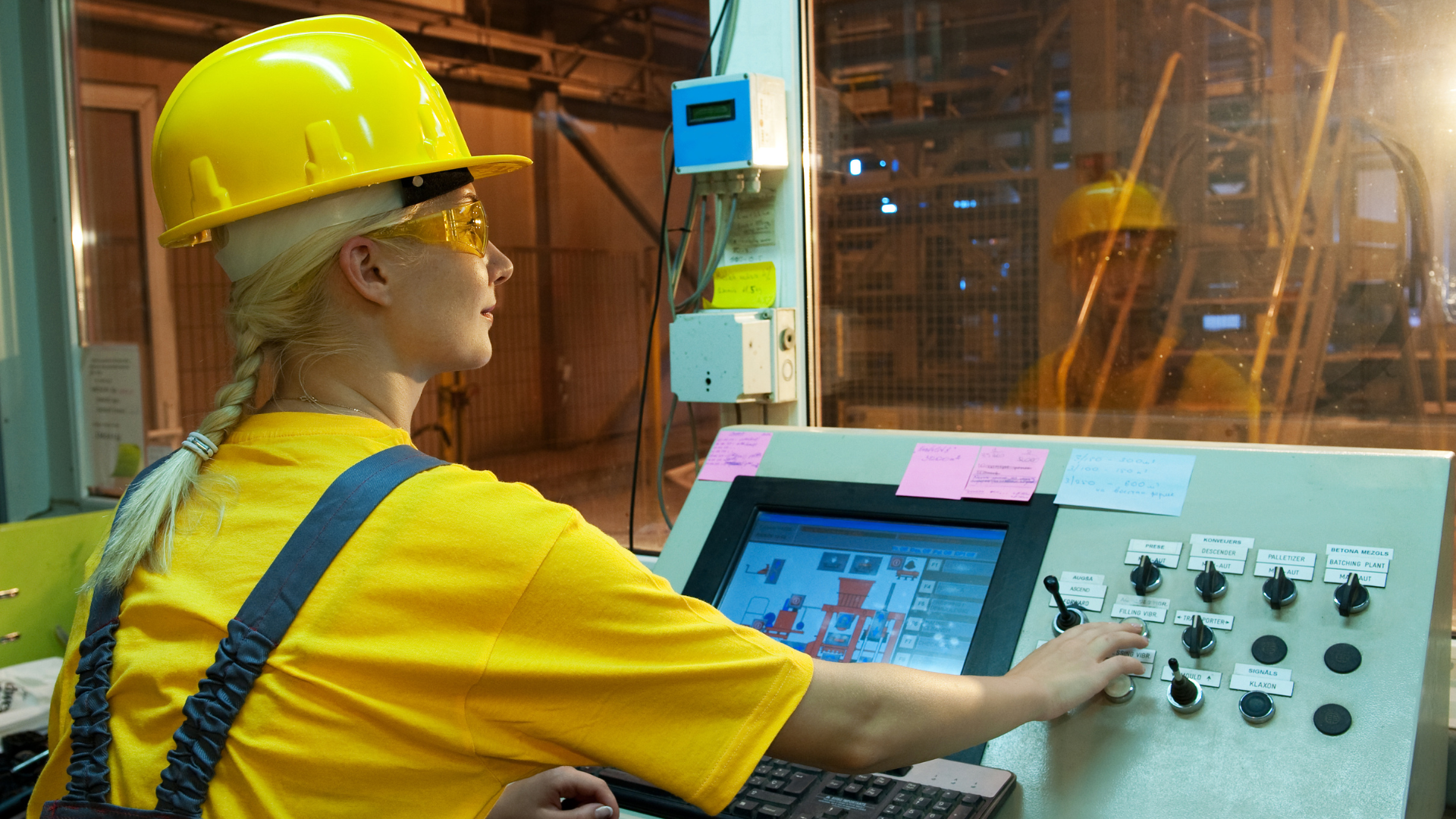 En kvinnlig processoperatör i gul skyddshjälm och säkerhetsglasögon arbetar vid ett kontrollbord med pekskärm och flera reglage. Hon övervakar industriell utrustning genom ett fönster i en fabriksmiljö med avancerade maskiner. Bilden är framtagen med AI.