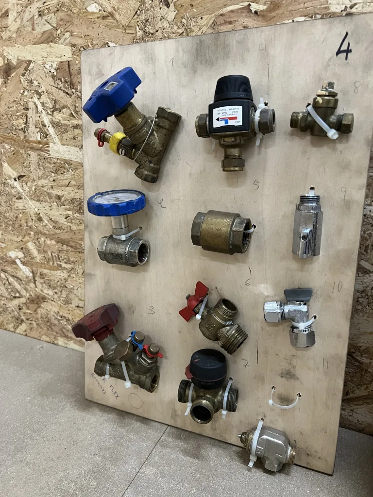 Träskiva med monterade ventiler och kopplingar i metall, uppmärkta med siffror för undervisning eller övning i VVS-teknik.