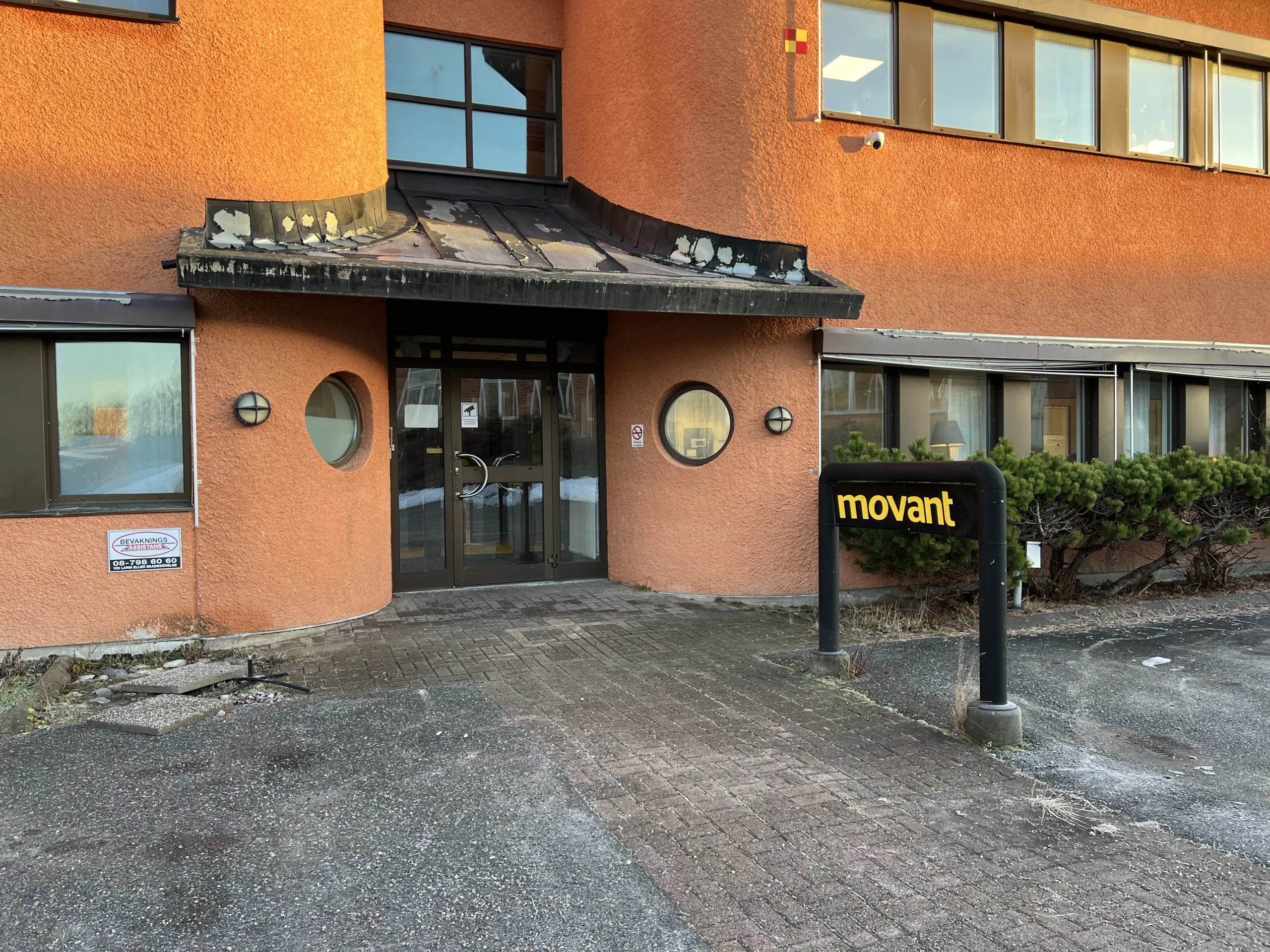 Entré till Movants byggnad med orange fasad, glasdörrar och en svart skylt med gul text som visar skolans namn.