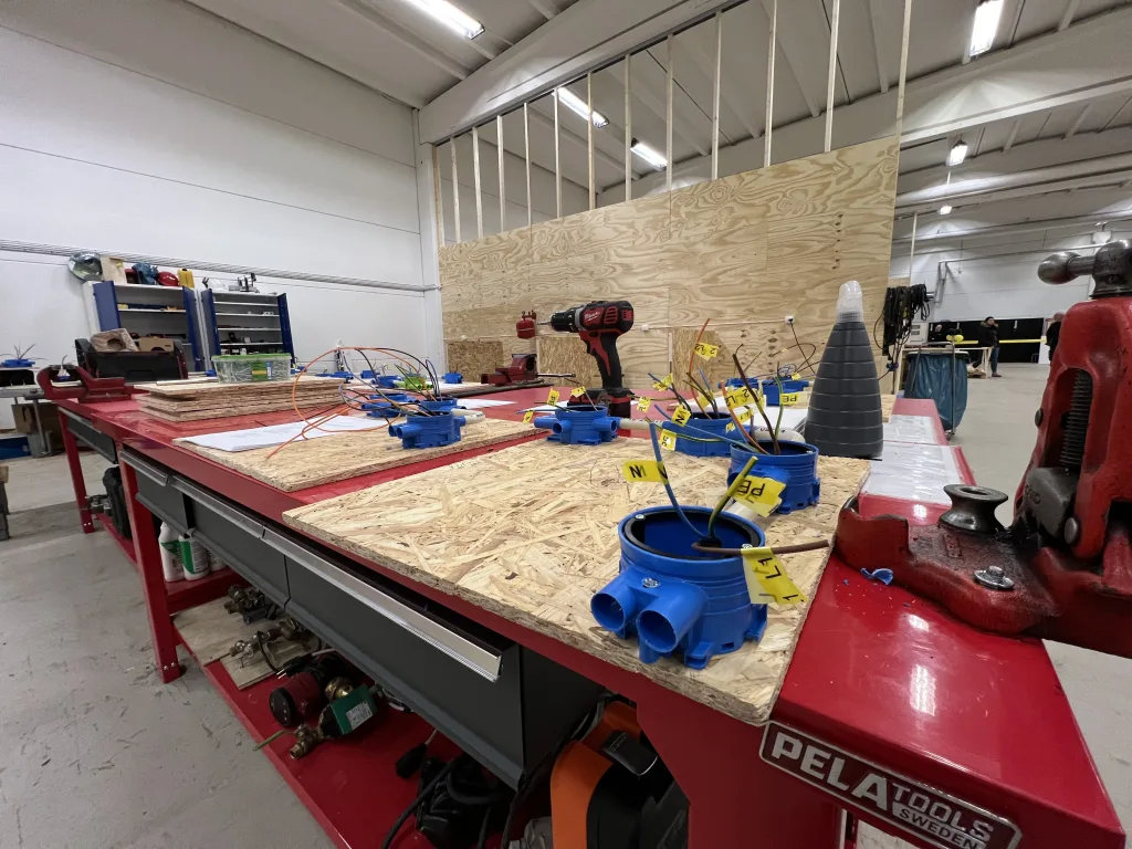 Arbetsbänk med elinstallationsmaterial monterat på träskivor, märkta ledningar och verktyg i en ljus verkstadsmiljö.