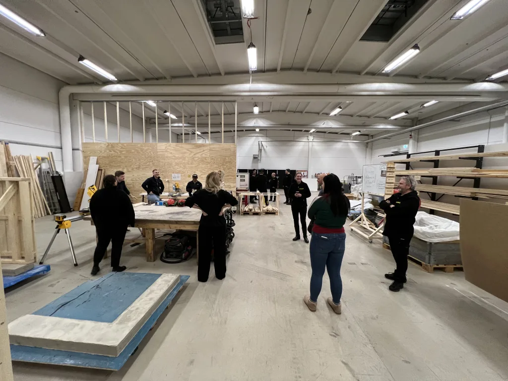 Större verkstadshall med grupper av människor samlade kring arbetsstationer i samtal och instruktion, omgiven av trämaterial och verktyg.