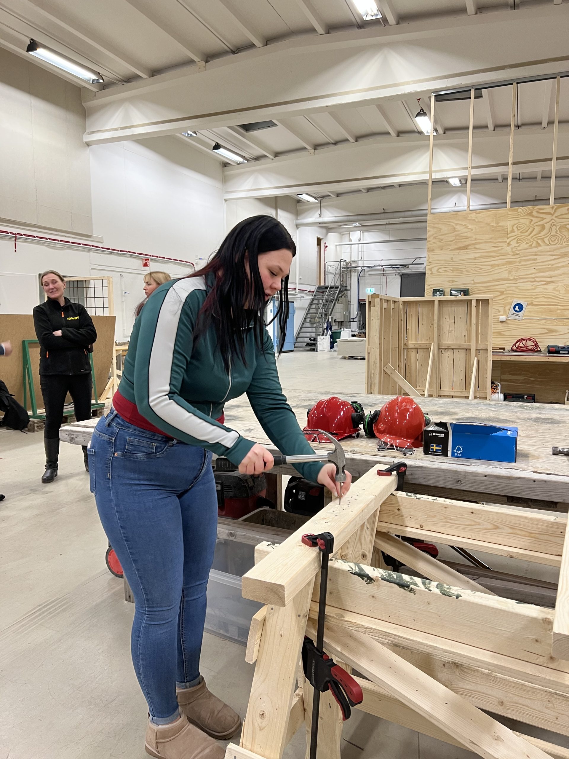Kvinna spikar i en träregel i en verkstad, med fokus på arbetet och skyddshjälmar liggande på bordet bakom henne.