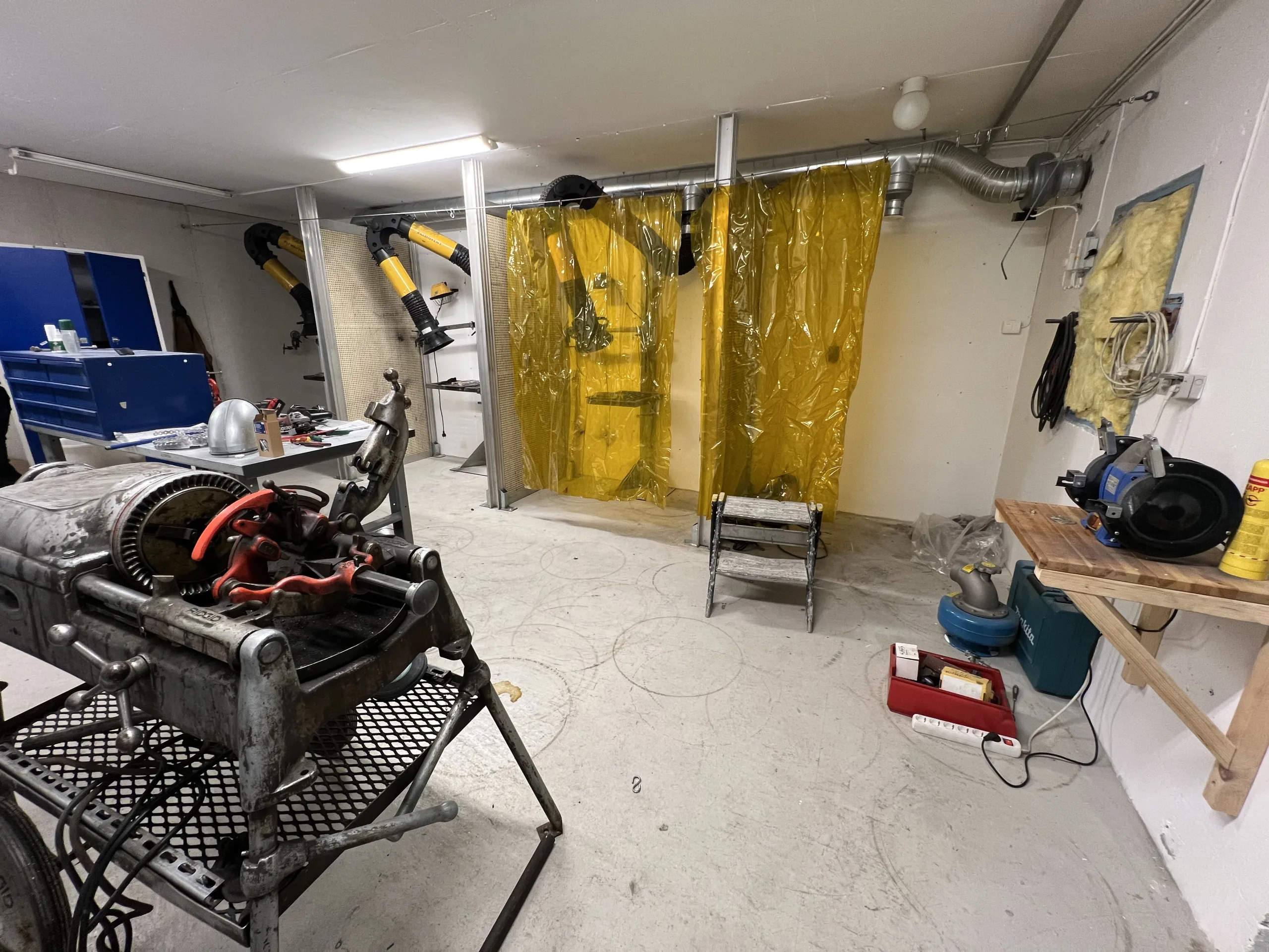 Verkstadsmiljö med svetsbås bakom gula plastdraperier, punktutsug med gula slangar, arbetsbänkar och verktyg utspridda i rummet.