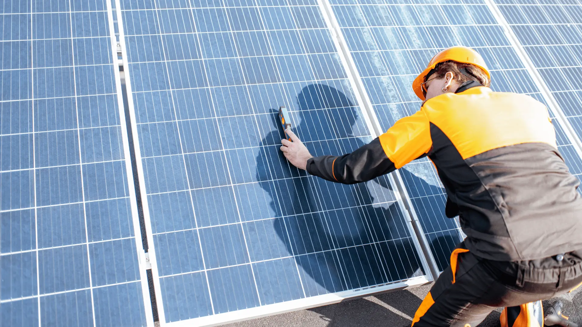 En solcellsmontör iförd orange och svart arbetsklädsel samt skyddshjälm inspekterar noggrant solpaneler med ett mätinstrument. Bilden visar den tekniska precision och säkerhetsmedvetenhet som krävs i yrket som solcellsmontör inom solenergiinstallationer.