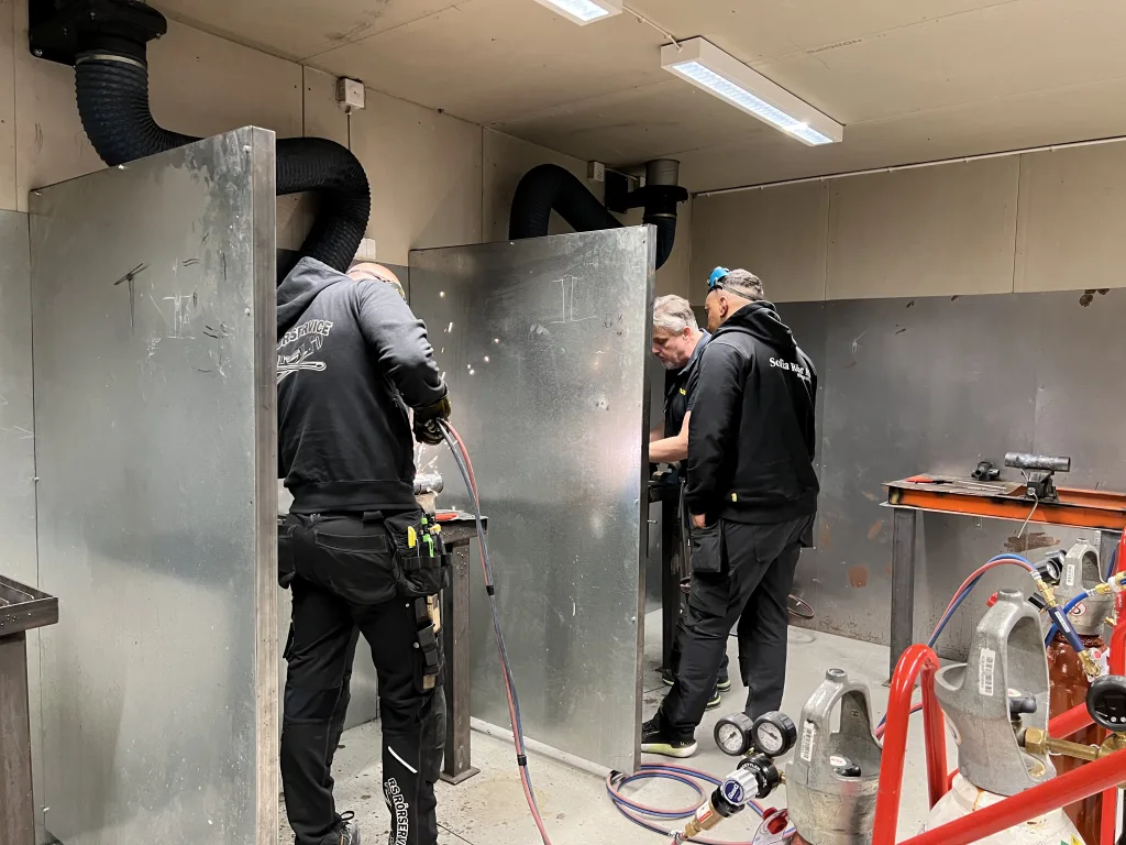 Tre personer arbetar i svetshörnor, en instruerar medan två andra svetsar rör med gnistor i luften.