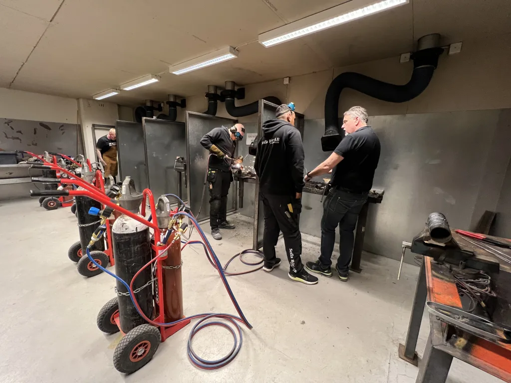 Flera personer arbetar vid svetshörnor med gassvetsning, handledning pågår och gasflaskor står på rad längs golvet.