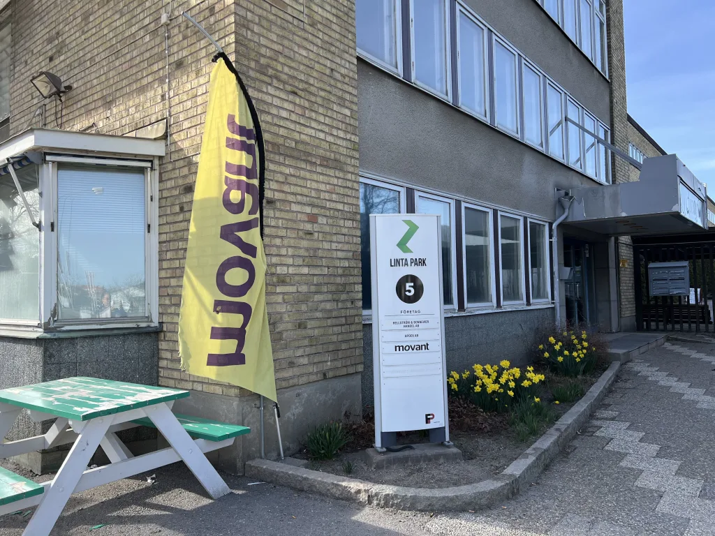 Fasaden till Movant i Stockholm med skyltar, gul flagga och vårblommor utanför entrén vid Linta Park.