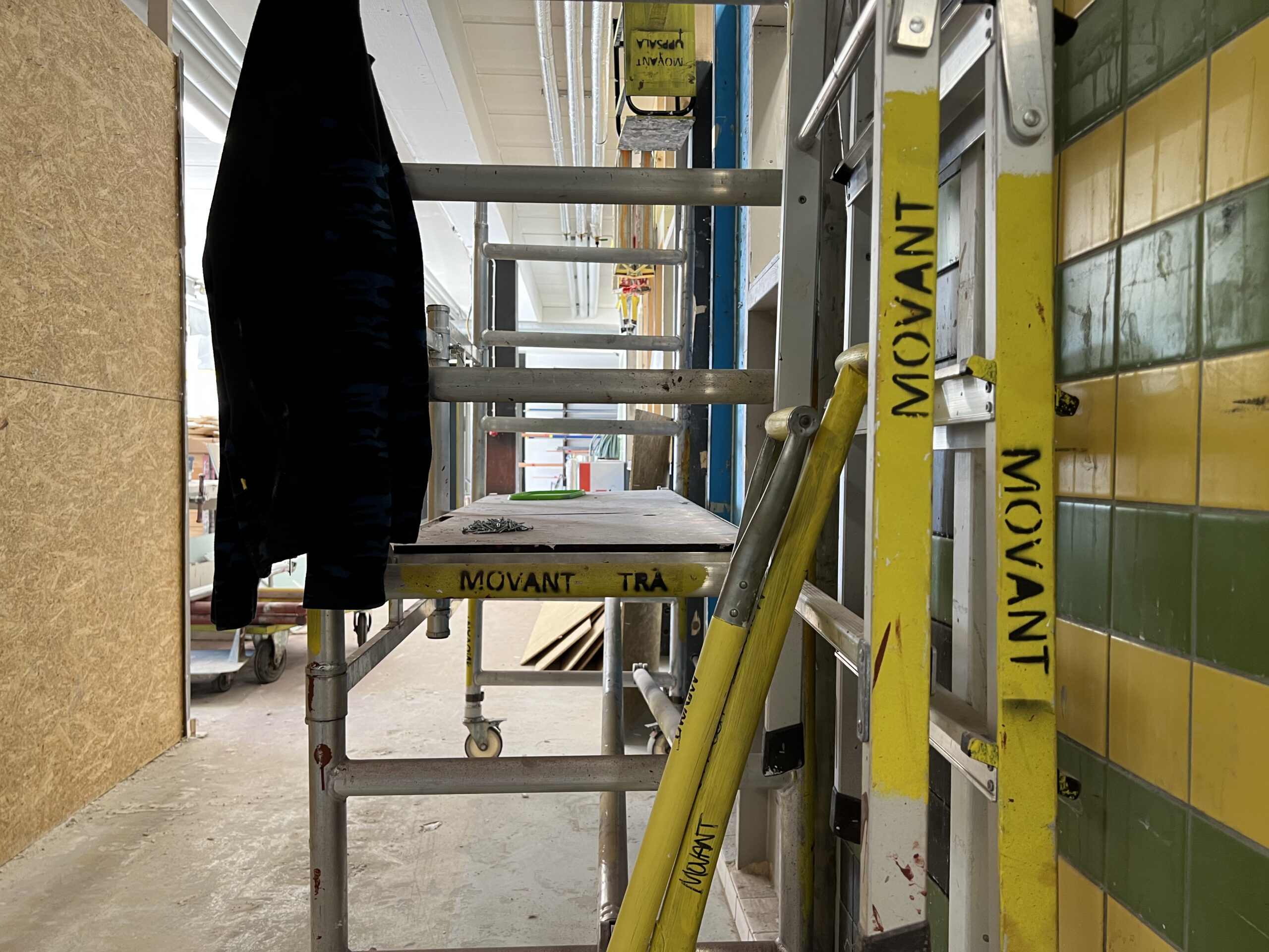 Rullställning och stegar märkta med “MOVANT” står uppställda i en gång mellan verkstadsrum med kaklade väggar och träpanel.