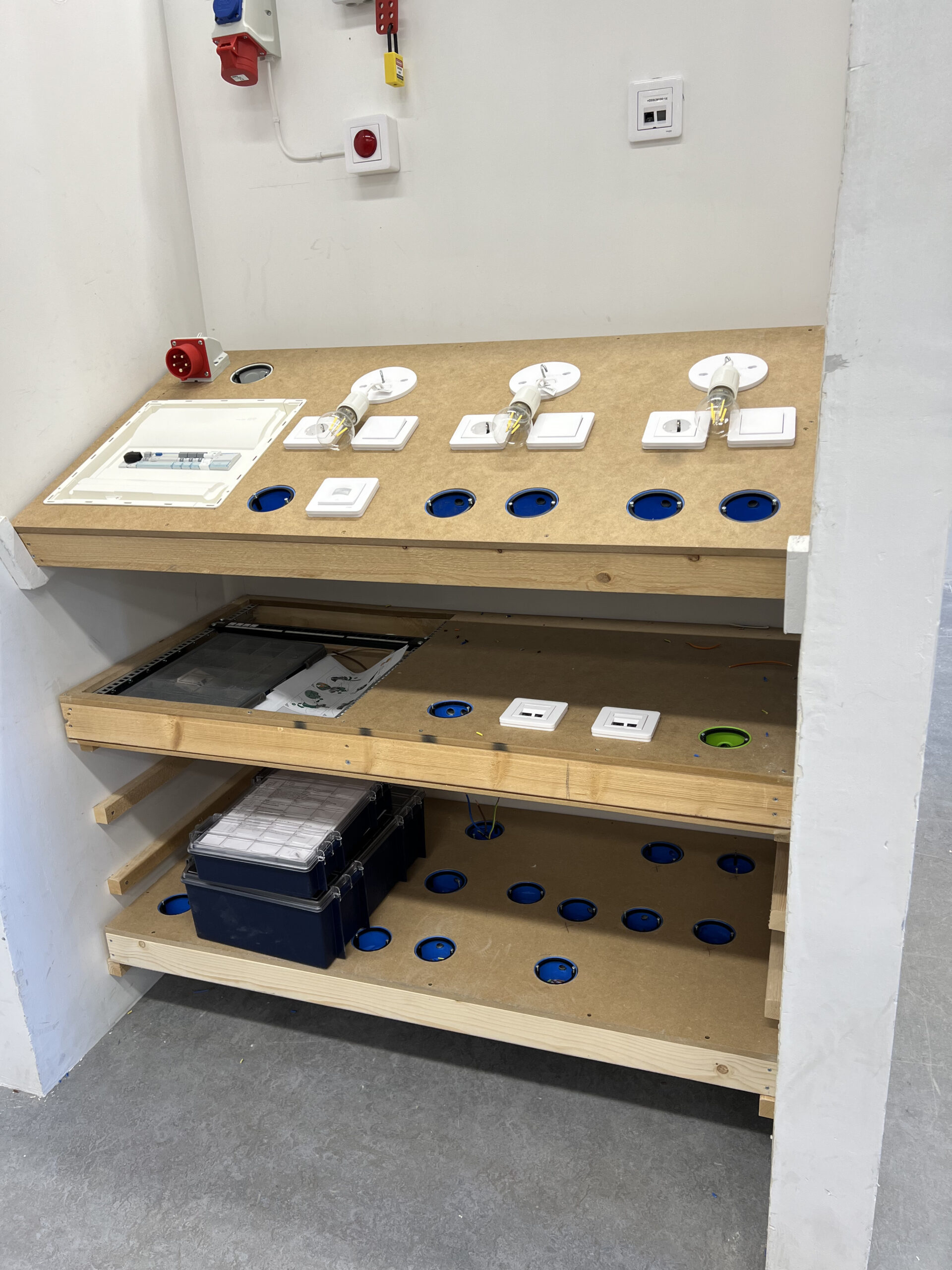 Närbild på en elinstallationsstation med flera strömbrytare, lampfattningar och elkomponenter. Verktygslådor och reservdelar är placerade på hyllor under arbetsytan.