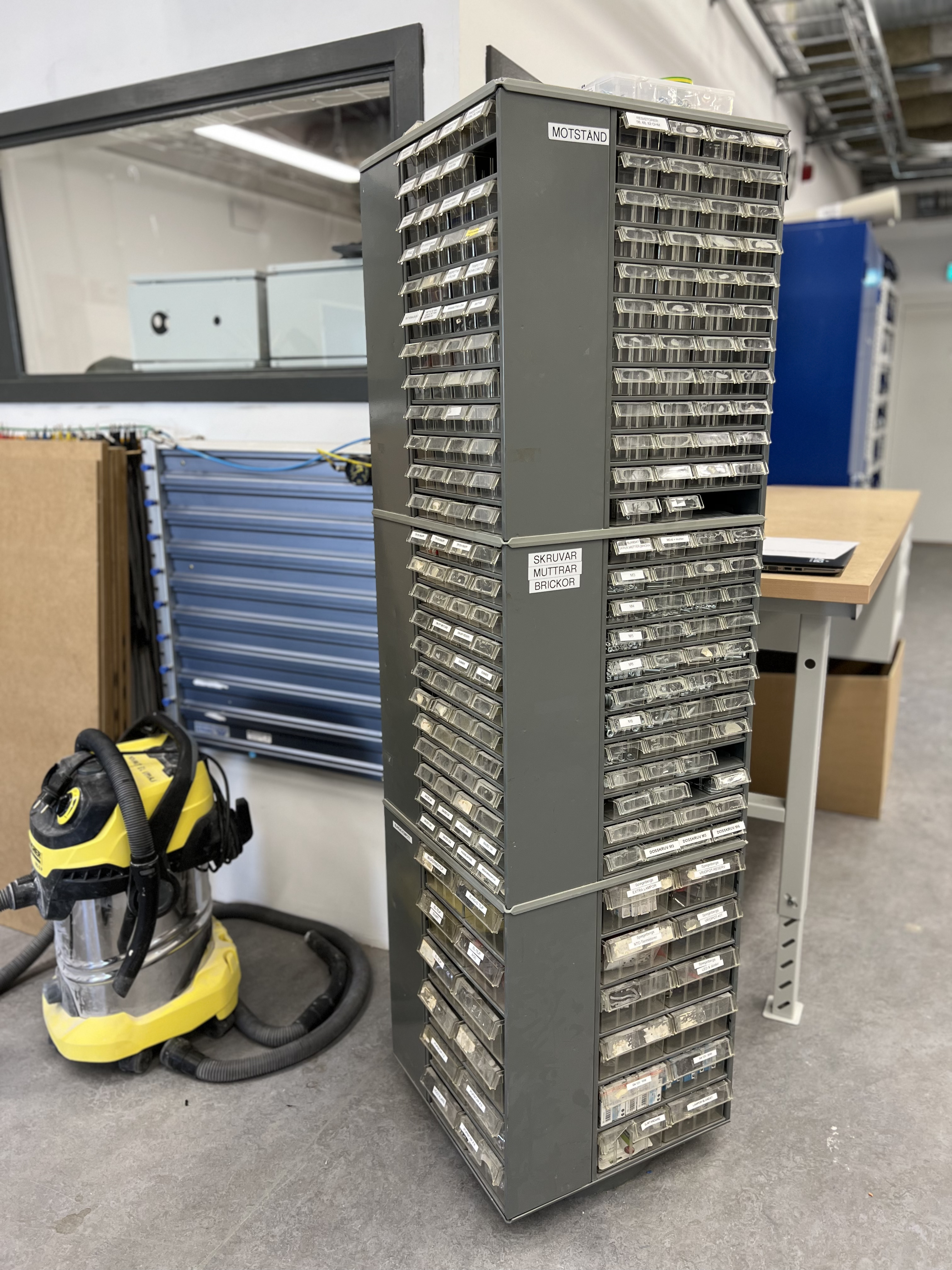 Sorteringsskåp för elektronikkomponenter med märkta lådor. Skåpet är märkt med etiketter som ”MOTSTÅND” och ”SKRUVAR MUTTRAR BRICKOR”.
