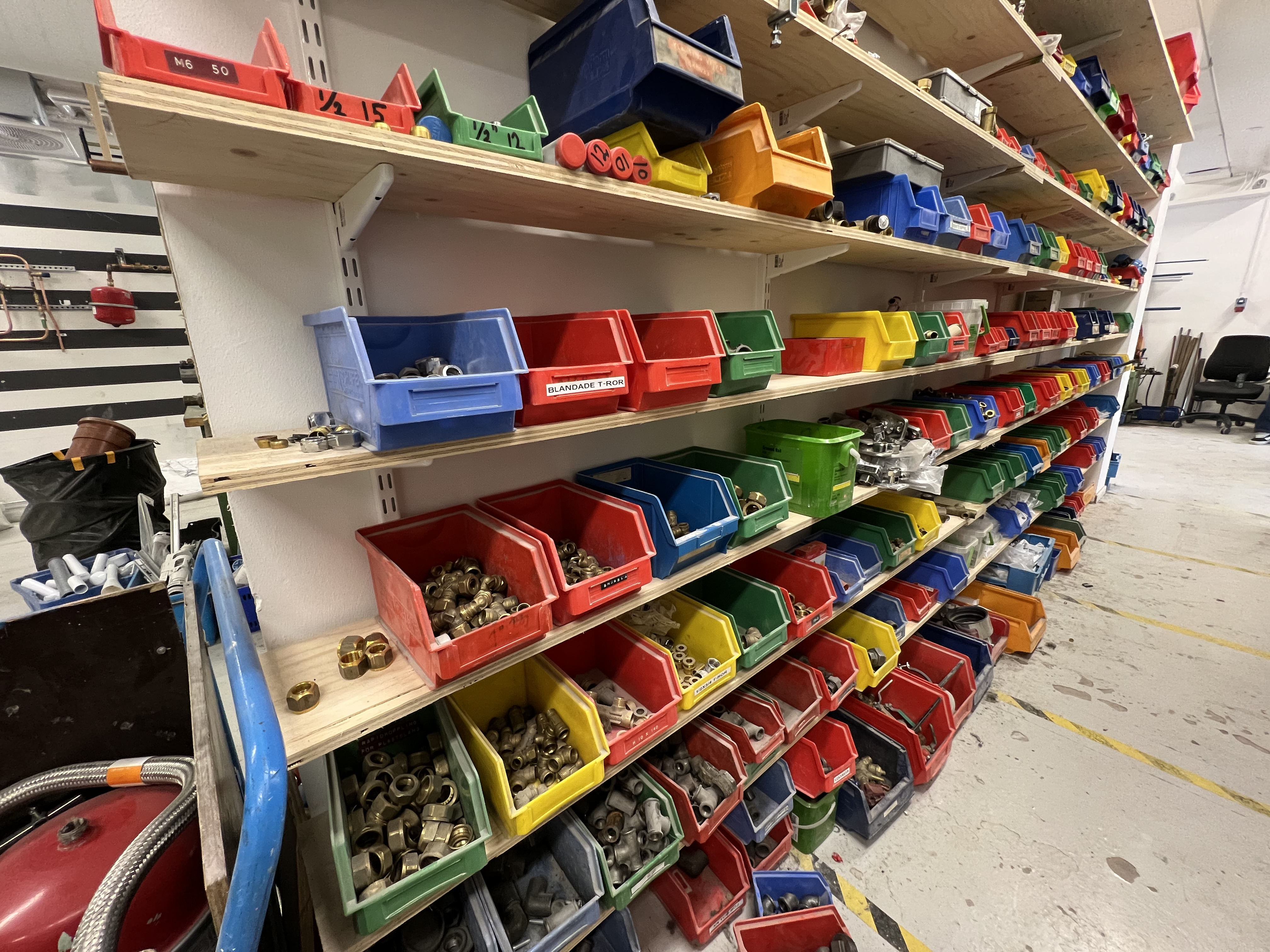Färgglada plastbackar fyllda med rördelar, muttrar och kopplingar står uppradade på hyllor i ett lagerutrymme i en verkstad.
