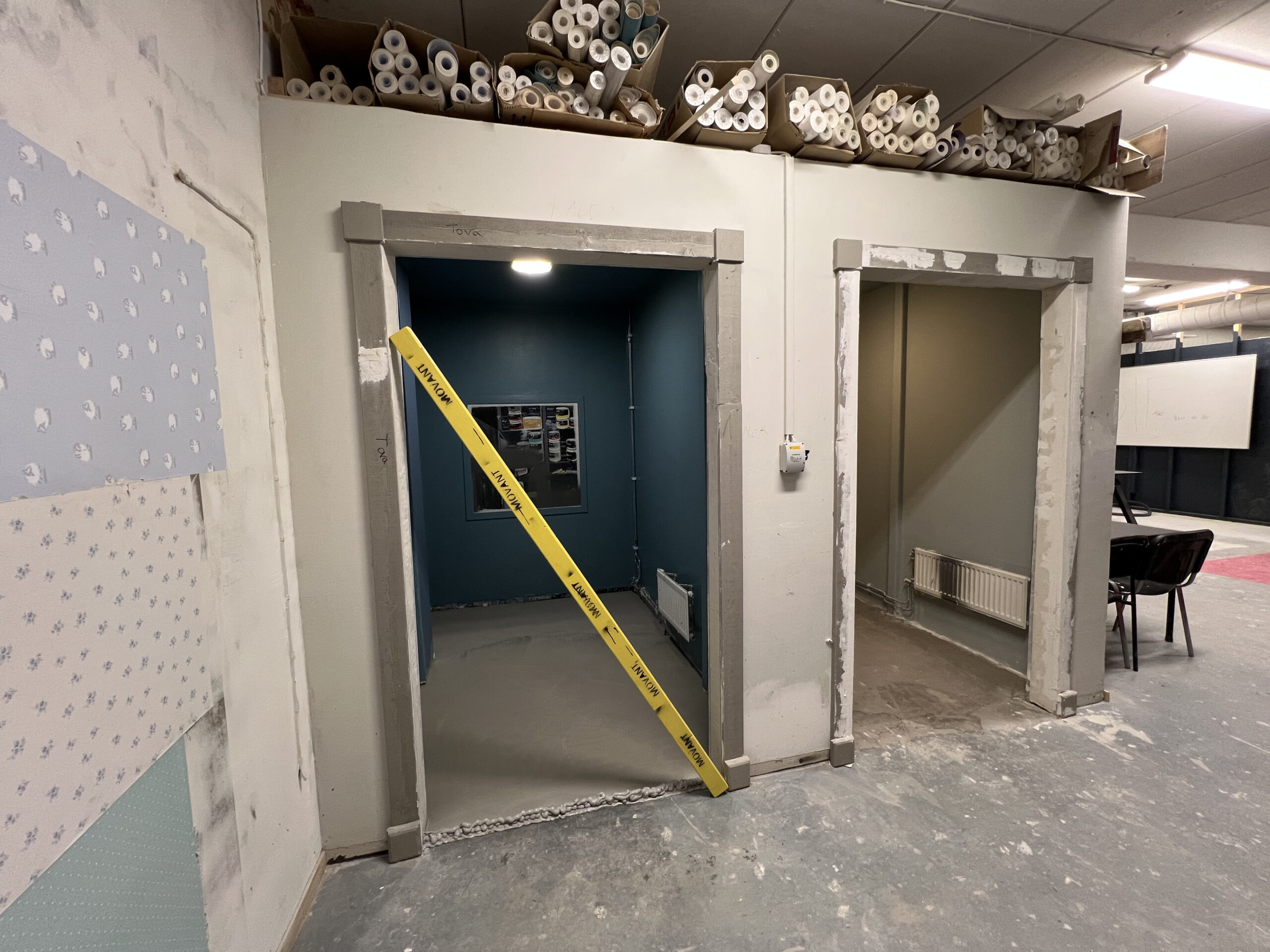 Två små rum i en utbildningslokal med väggar i olika färger, golv under renovering och tapetprover monterade på väggen till vänster.
