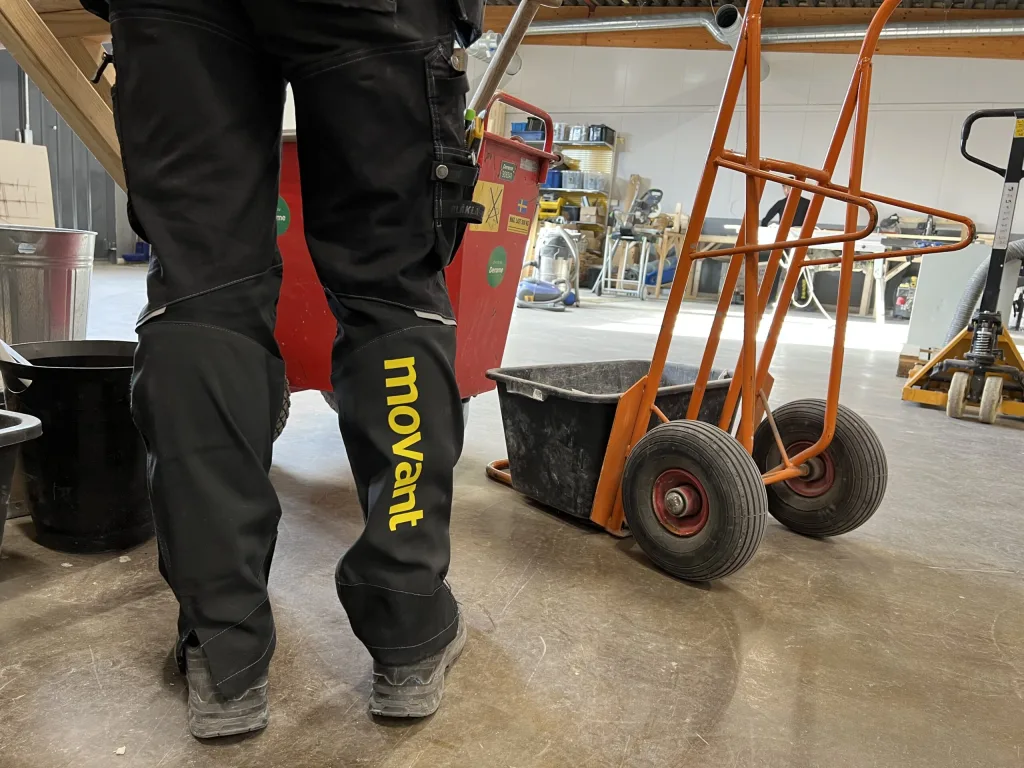 Elev i arbetsbyxor med Movant-logga i verkstadsmiljö med verktyg och transportvagn.