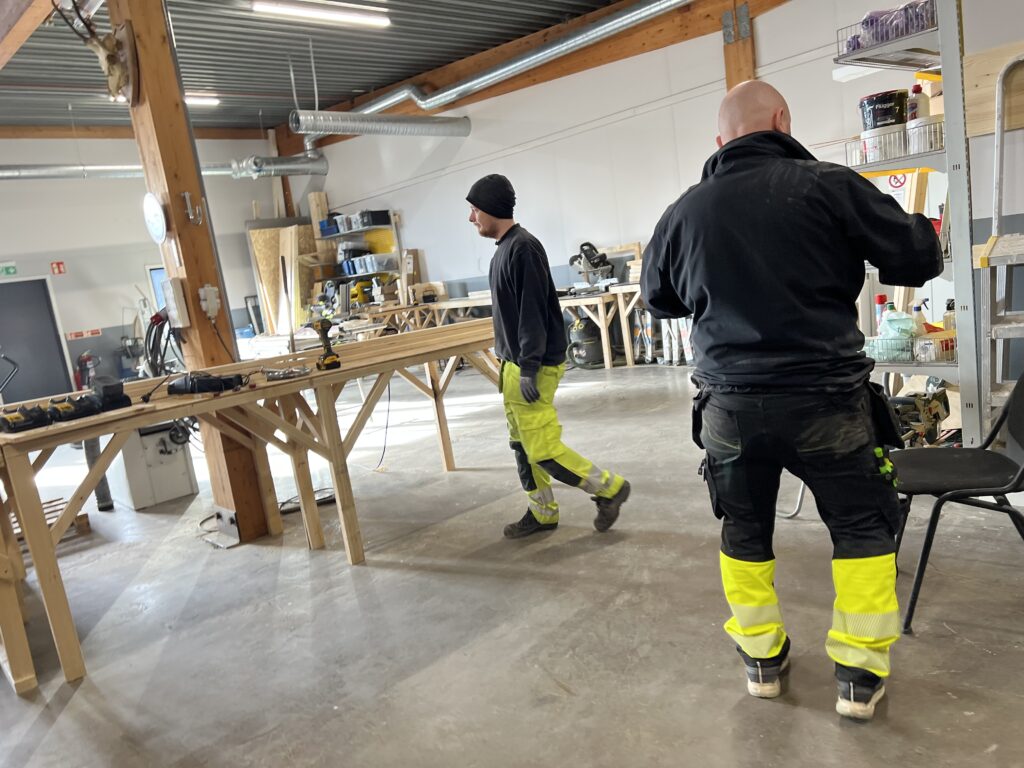 Två personer i arbete vid träbänkar i Movant Varbergs bygglokaler med verktyg i fokus.