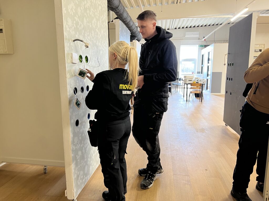 Två elever arbetar med elinstallation i en utbildningsmiljö på Movant Varberg. Eleven i förgrunden bär tröja med texten ”Movant Elektriker”.