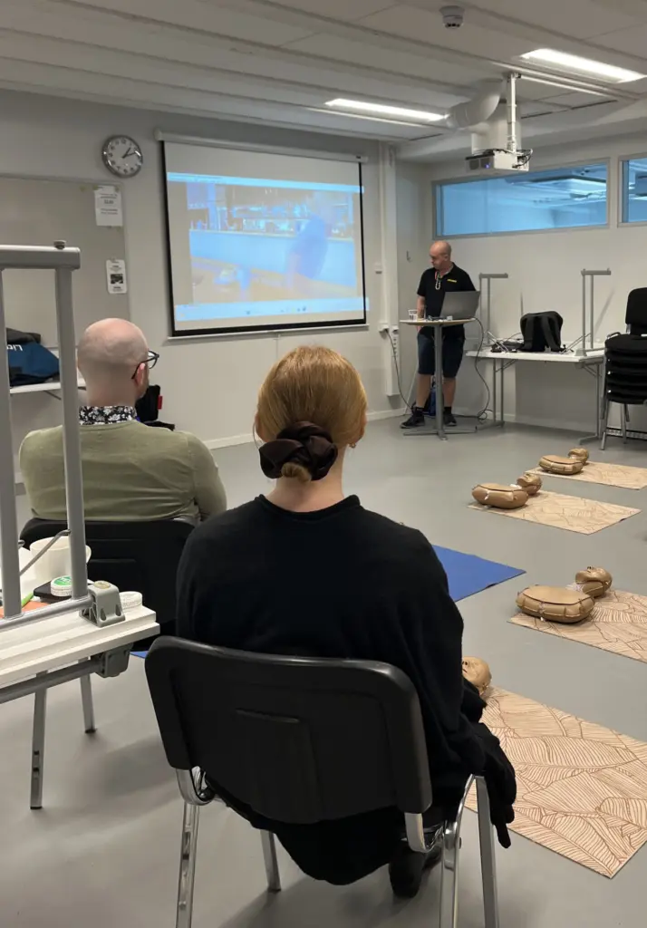 Deltagare sitter i ett klassrum under en HLR-utbildning hos Movant. En utbildare står längst fram och håller genomgång medan en film visas på projektor. På golvet ligger övningsdockor redo för praktiska moment.
