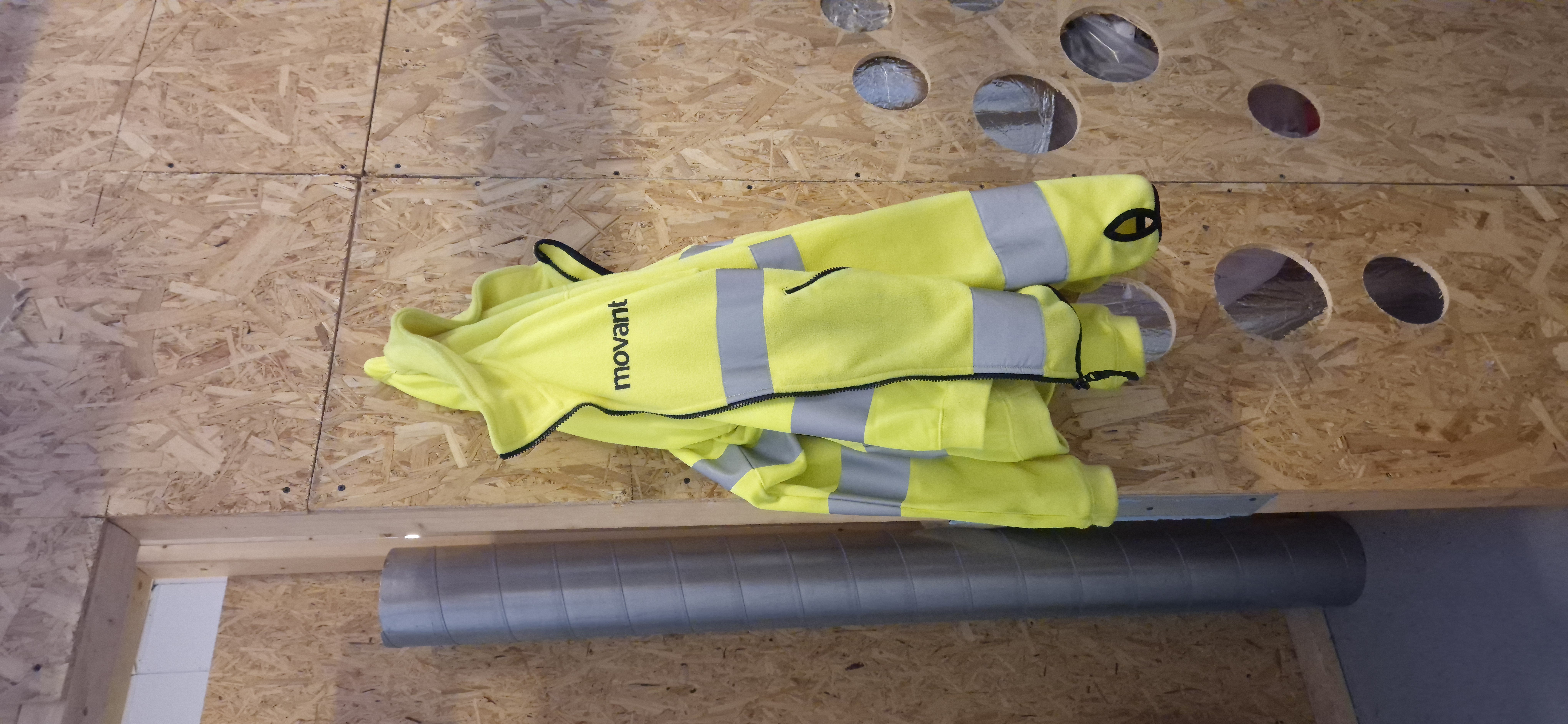 Reflexjacka från Movant i verkstad Stockholm fotograferad i en industriell utbildningsmiljö med träväggar och ventilationsrör. Bilden visar arbetskläder för bygg och hantverk med fokus på säkerhet, synlighet och professionell arbetsmiljö för elever och yrkesverksamma.