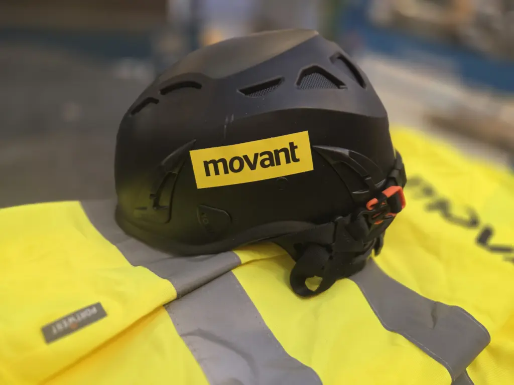 Närbild på svart skyddshjälm med Movant-logotyp placerad på gul reflexjacka i verkstad Stockholm. Bilden symboliserar säker arbetsmiljö, yrkesutbildning och professionell skyddsutrustning inom bygg, industri och praktiskt arbete i modern verkstadsmiljö.